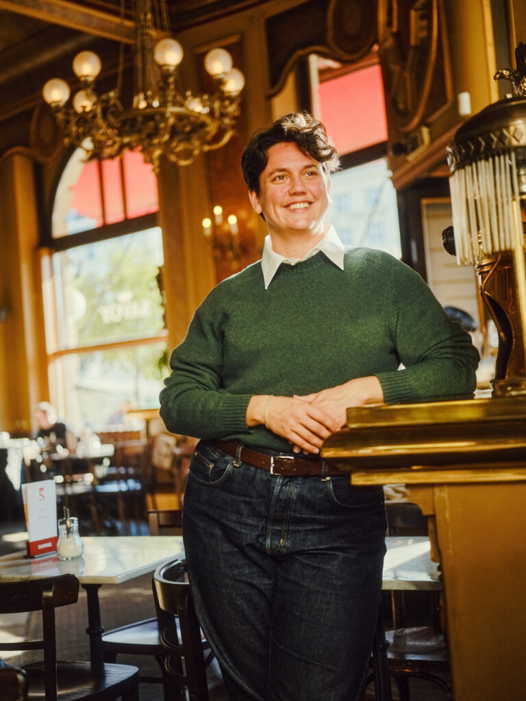 Clar Gallistl lehnt an einem Tisch. Clar trägt kurzes, welliges Haar, einen grünen Pullover und ein Hemd darunter. Clar lächelt.