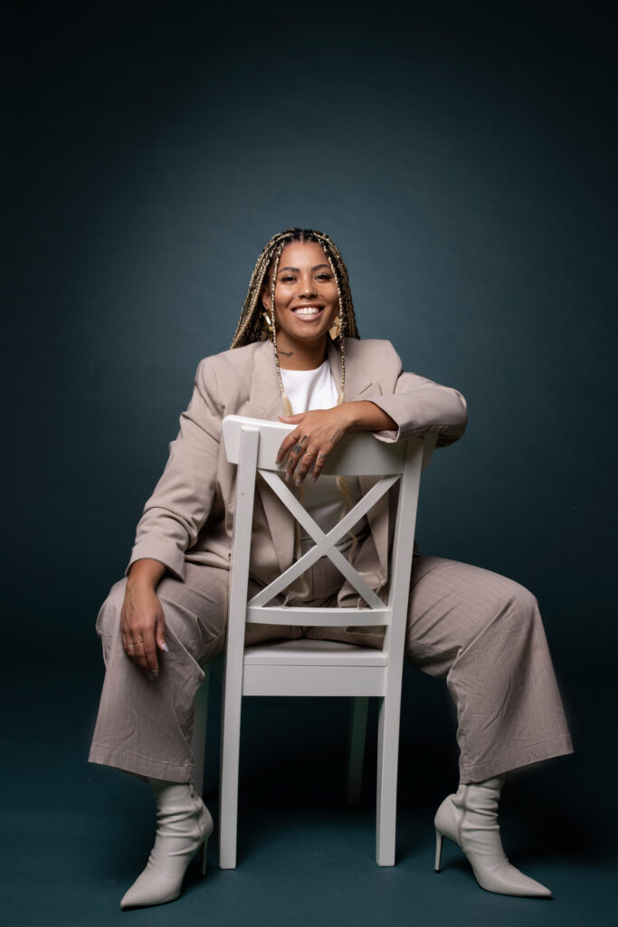 Lächelnde Frau mit langen, geflochtenen Haaren sitzt locker auf einem weißen Stuhl vor einem dunklen, blaugrünen Hintergrund. Sie trägt einen hellbeigen Hosenanzug mit weißem Top und weißen Stiefeletten. Mit einem Arm lehnt sie über die Rückenlehne des Stuhls und blickt in die Kamera.