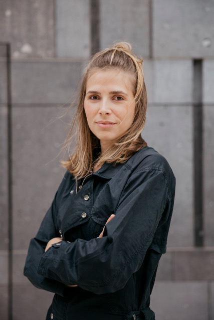  Porträt einer Frau mit blondem, halb hochgebundenem Haar, die mit verschränkten Armen vor einer grauen Betonwand steht. Sie trägt eine dunkelgraue Jacke und blickt direkt in die Kamera; ihr Gesicht wirkt ruhig und selbstbewusst, leichter Wind bewegt einzelne Haarsträhnen.